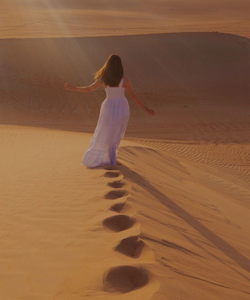 Person in a white dress walking on sand dunes with footprints leading the way.