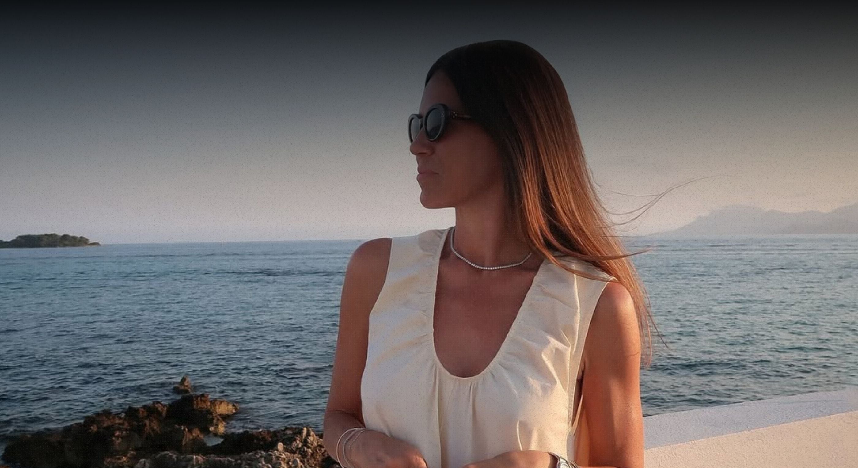 Woman in a white sleeveless top standing by the ocean with sunglasses on.