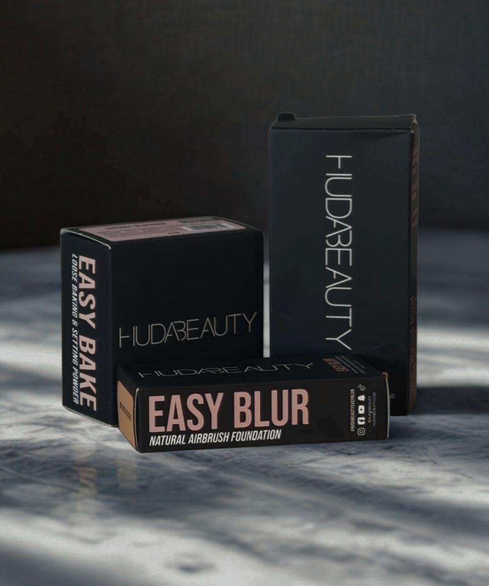Huda Beauty makeup products including 'Easy Bake' and 'Easy Blur' on a dark surface.