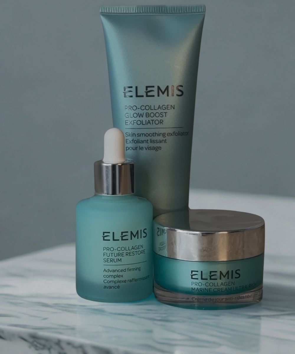 Elemis skincare products including a serum, exfoliator, and cream on a marble surface with a gray background.