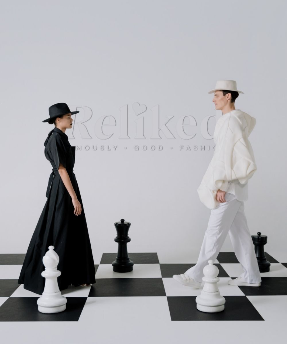 Two people standing on a chessboard with large chess pieces, wearing black and white outfits.