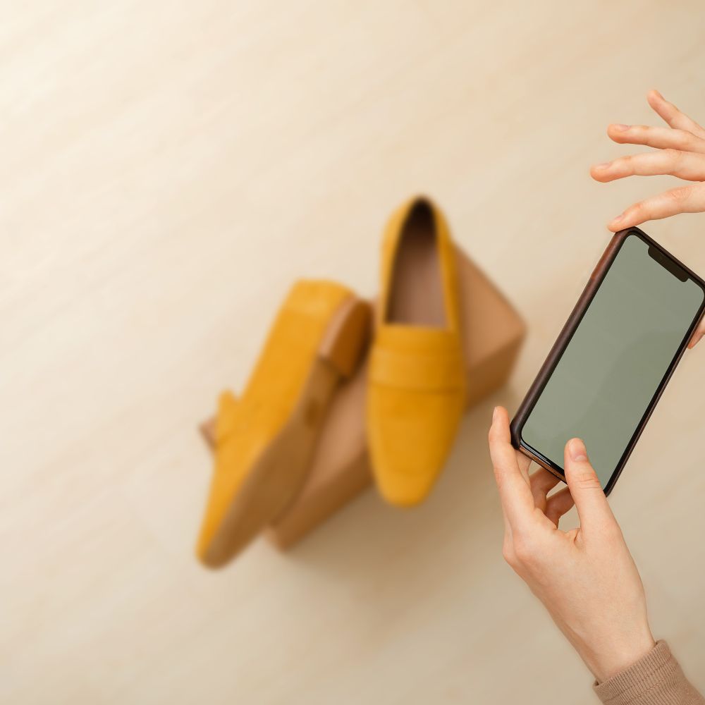 Yellow loafers on a beige surface with a hand holding a phone.