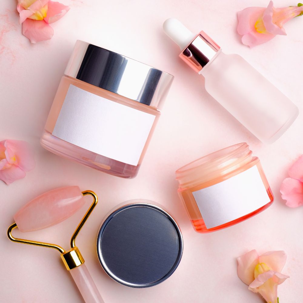 Set of skincare products including a jar, dropper bottle, roller, and compact on a pink background with flowers.