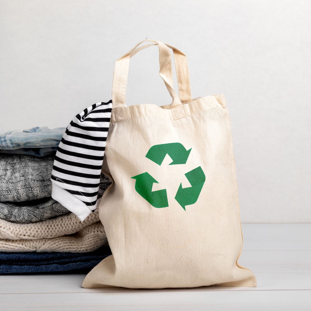 Beige tote bag with green recycling symbol next to folded clothes on a light wooden surface